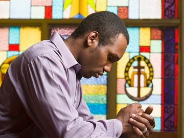 African American man praying in church with stained glass in background